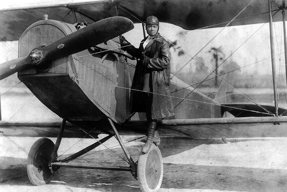 Bessie Coleman, an early airshow pilot, poses on the wheel of her stunt biplane. She is a Black woman in a long, dark dress wearing a leather helmet and a proud, serious expression.