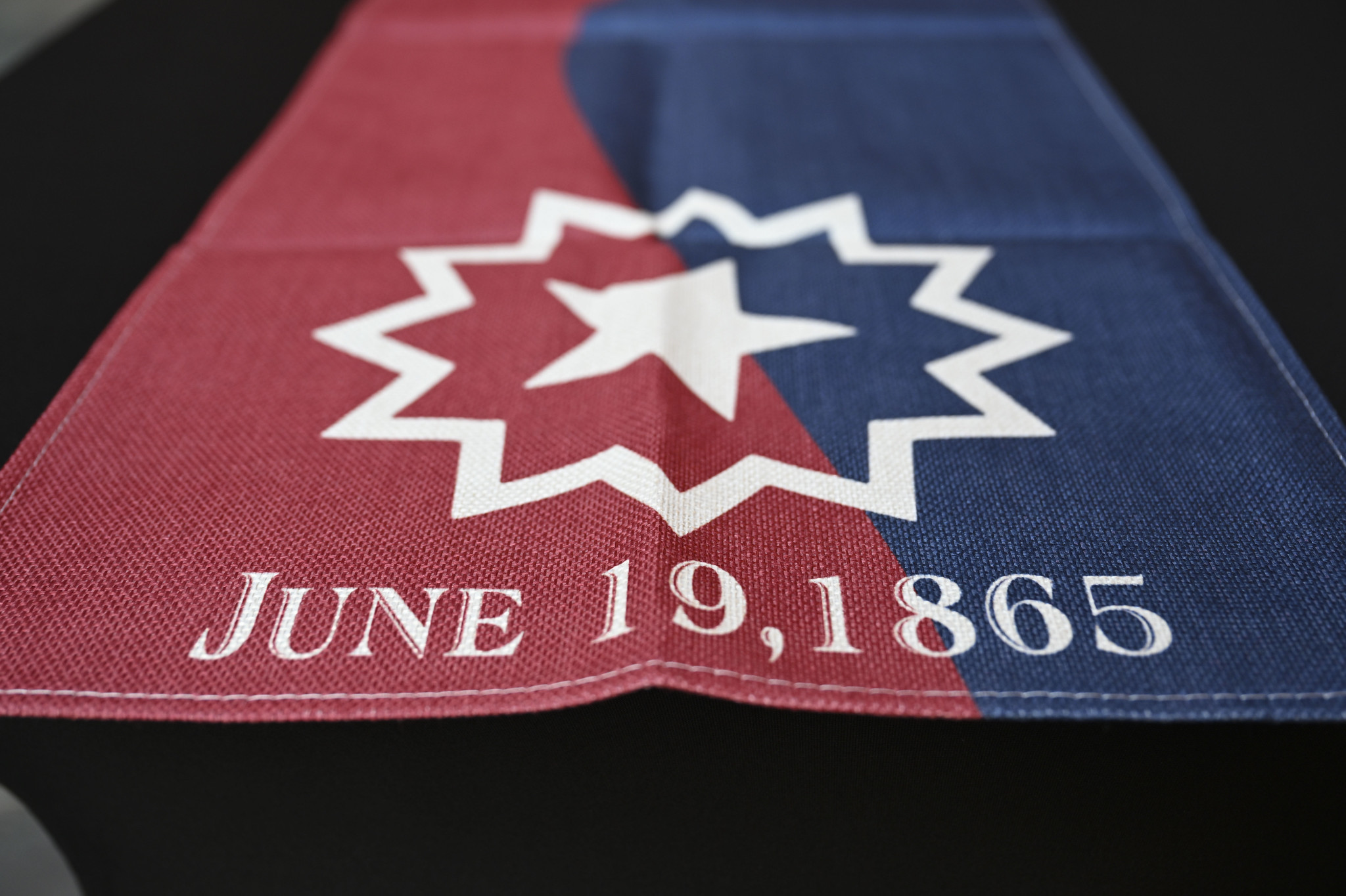 Juneteeth flag, red on the left, dark blue on the right, with a white five-pointed star surrounded by a twelve-pointed sunburst above white text reading June 19, 1865