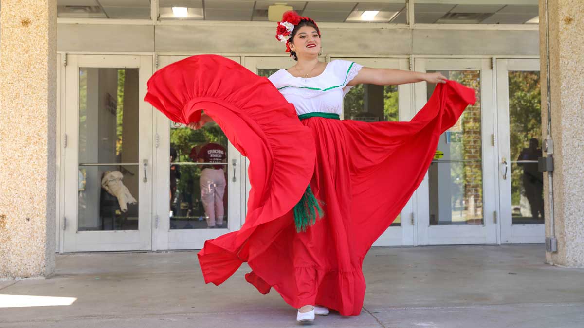 a dancer in traditional mexican costume, with a full red skirt and white blouse, twirls in front of the entrance to the student center