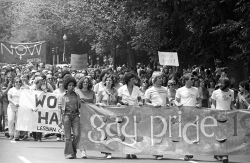 photograph of a diverse crowd of people, mostly men, walking down a tree-lined street carrying a banner reading "gay pride 1970"